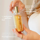 Hand hält Golden-Glow-Oil, andere Hand öffnet die Flasche, mit Informationstext.