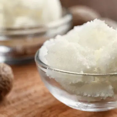 Glasschale mit Sheabutter steht auf einem Holztisch, daneben unscharfe Nüsse.
