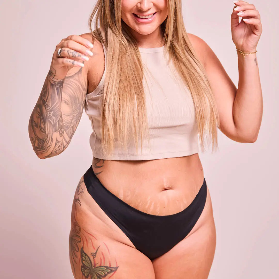 Laughing blonde woman with a tattooed arm in a beige crop top and the black Seamless String